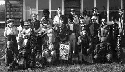 Old photograph of a large group of people in fancy dress outside a wooden hut, with a drum and a banner